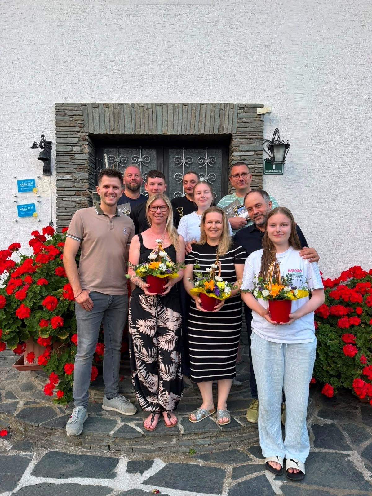 Eine Gruppe von Menschen, sowohl Männer als auch Frauen, stehen vor einem Gebäude mit Blumen in den Händen. Sie lächeln und posieren für ein Foto.