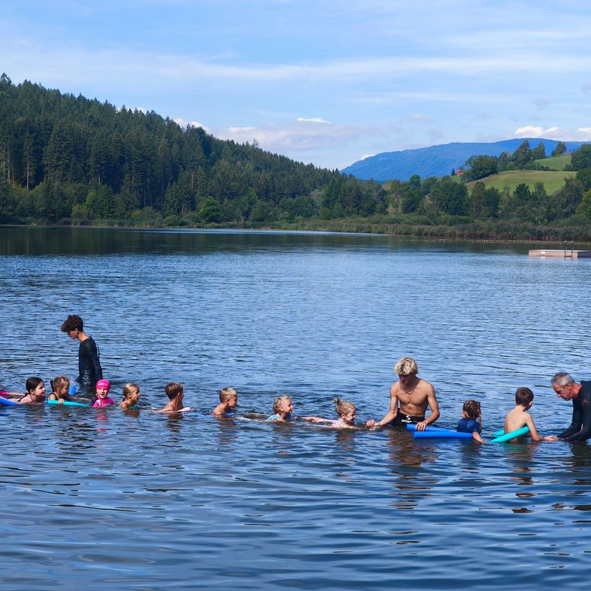 Eine Gruppe von Kindern und Erwachsenen schwimmt in einem See mit Bergen im Hintergrund.