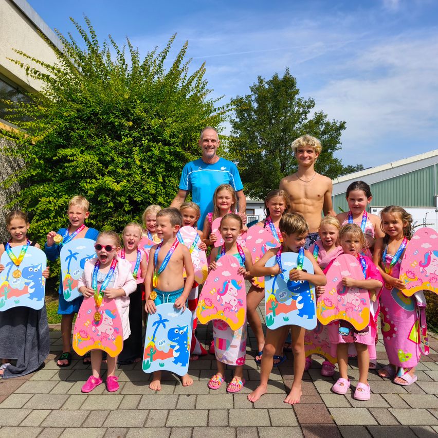Eine Gruppe junger Schwimmer, geschmückt mit bunten Handtüchern und Medaillen, steht zusammen mit ihrem Trainer und posiert für ein Foto.
