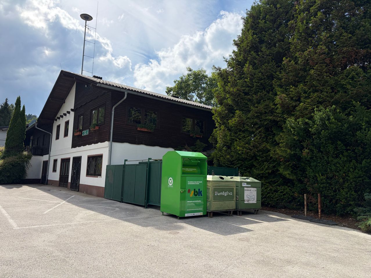 Ein moderner grüner Recyclingbehälter steht vor einem Gebäude mit braunen und weißen Wänden. Hinter dem Behälter befinden sich zwei weitere grüne Behälter. Das Gebäude hat einen Turm und mehrere Fenster.