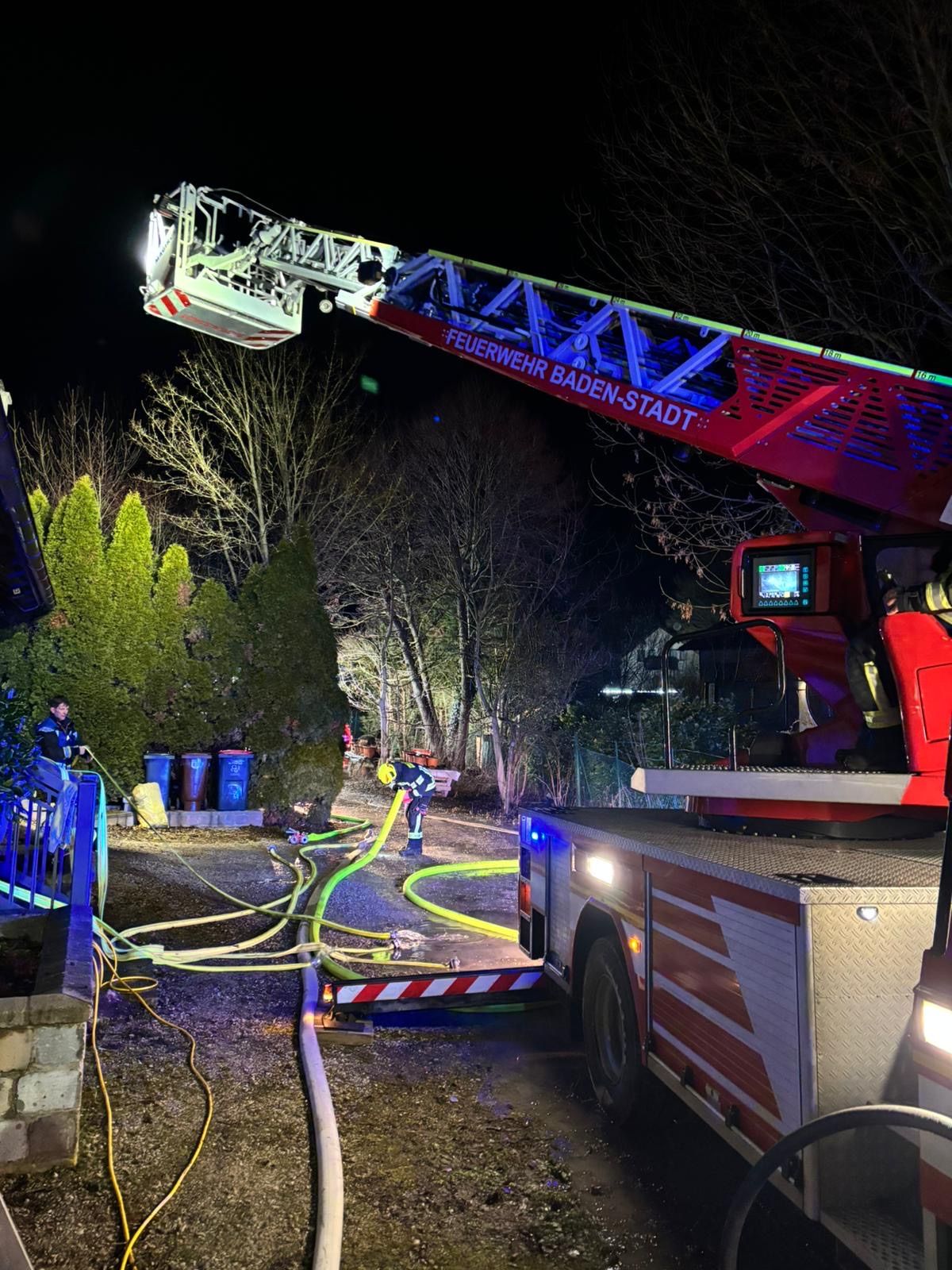 Ein Feuerwehrfahrzeug ist nachts geparkt, mit ausgeklappter Leiter. Ein Feuerwehrmann ist auf der Leiter. Bäume und Mülleimer sind in der Nähe.