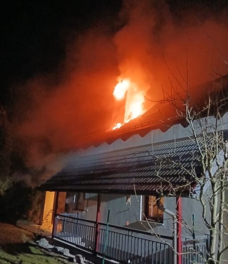 Ein Haus steht bei Nacht in Flammen, mit Feuer, das aus dem Dach schlägt. Rauch steigt aus dem Brand auf.