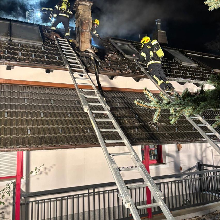 Drei Feuerwehrleute sind nachts auf dem Dach eines Gebäudes und klettern auf Leitern. Sie tragen gelbe Helme und gelbe und schwarze Uniformen. Rauch kommt aus dem Schornstein.