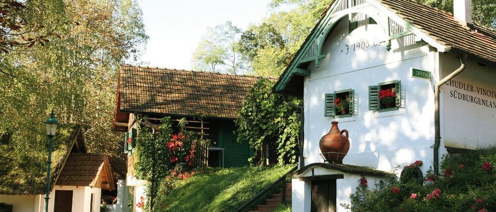 Ein traditionelles weißes Gebäude mit braunem Rand, einem braunen Dach und einem großen braunen Krug obenauf, mit Kletterpflanzen und Blumen davor.