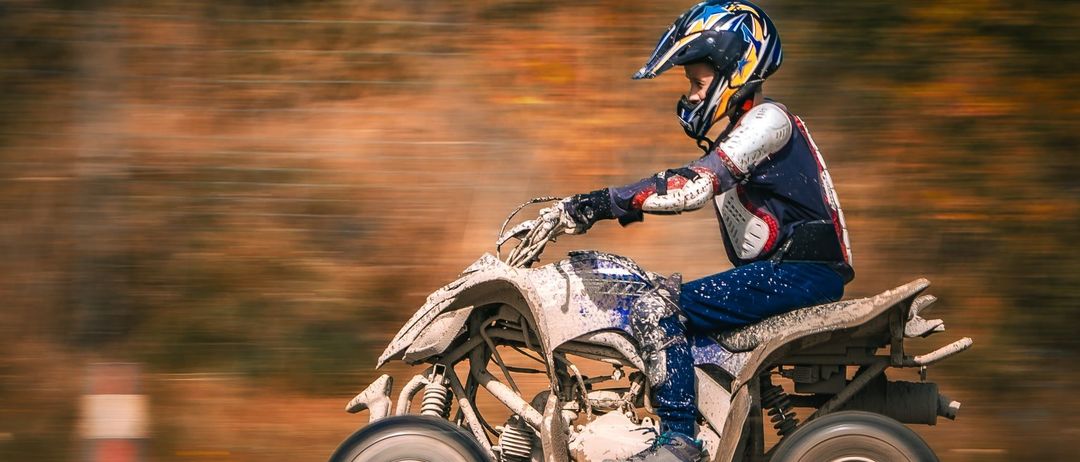 Eine Person fährt ein vierrädriges Motorrad im Wald. Der Fahrer trägt einen Helm und ist mit Schlamm bedeckt. Der Hintergrund ist verschwommen.