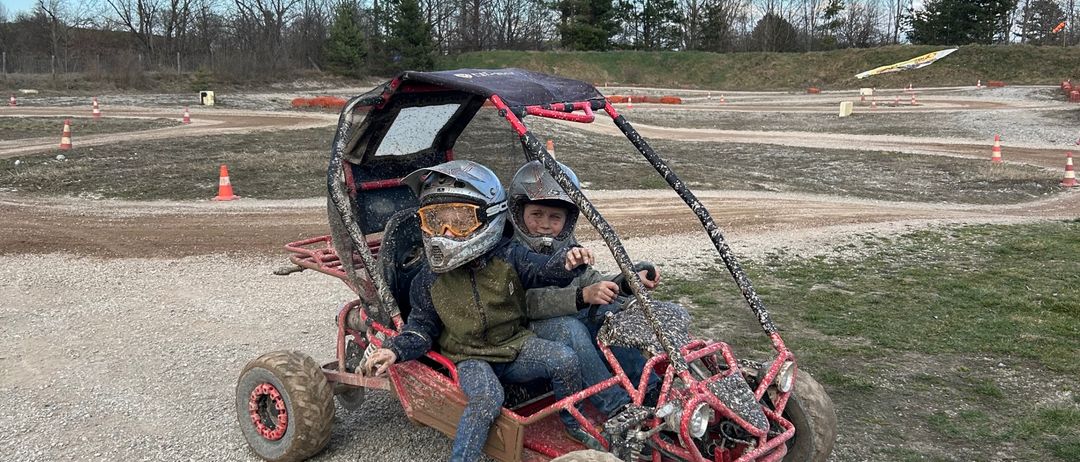 Zwei Kinder in Helmen fahren einen roten Go-Kart auf einer Kiesstrecke, umgeben von orangefarbenen Verkehrskegel unter blauem Himmel.