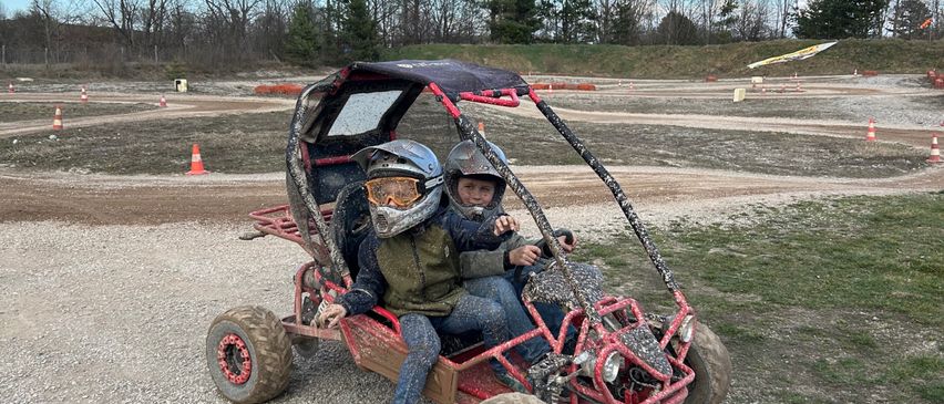 Zwei Kinder in Helmen fahren einen roten Go-Kart auf einer Kiesstrecke, umgeben von orangefarbenen Verkehrskegel unter blauem Himmel.