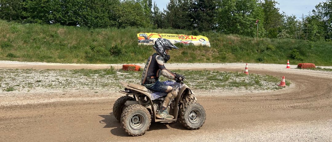Eine Person fährt ein Quad auf einer Schlammstrecke, mit orangefarbenen Verkehrskegel und einem Grashügel im Hintergrund. Der Himmel ist blau mit einigen Wolken.