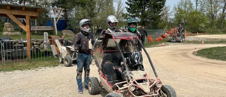 Drei Personen in Helmen schieben ein Go-Kart auf einer Erdstrecke, mit einer Holzstruktur und Bäumen im Hintergrund.
