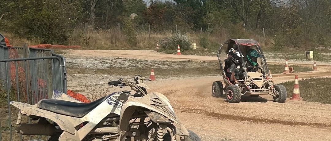 Zwei Geländefahrzeuge, eines mit vier Rädern und das andere ein zweirädriges Buggy, stehen auf einer unbefestigten Strecke mit Bäumen und Verkehrskegeln im Hintergrund.
