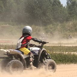 Ein junger Junge fährt ein Yamaha-ATV auf einer Dirt-Piste, umgeben von grüner Vegetation. Er trägt einen Helm und Schutzausrüstung. Andere Fahrer folgen dahinter.