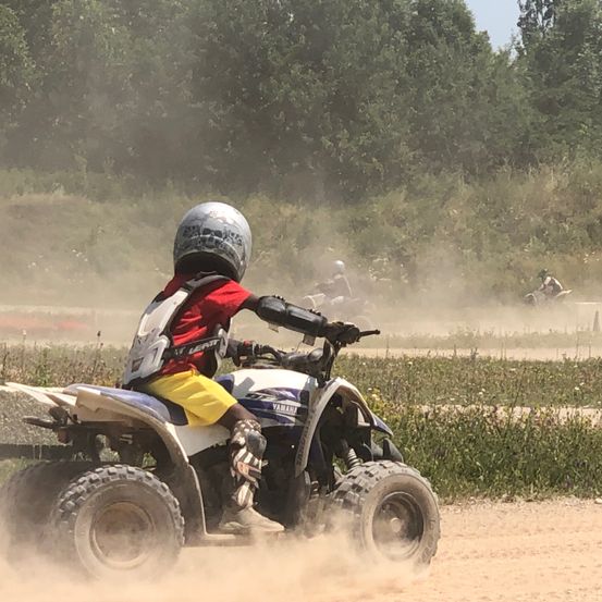 Ein junger Junge fährt ein Yamaha-ATV auf einer Dirt-Piste, umgeben von grüner Vegetation. Er trägt einen Helm und Schutzausrüstung. Andere Fahrer folgen dahinter.