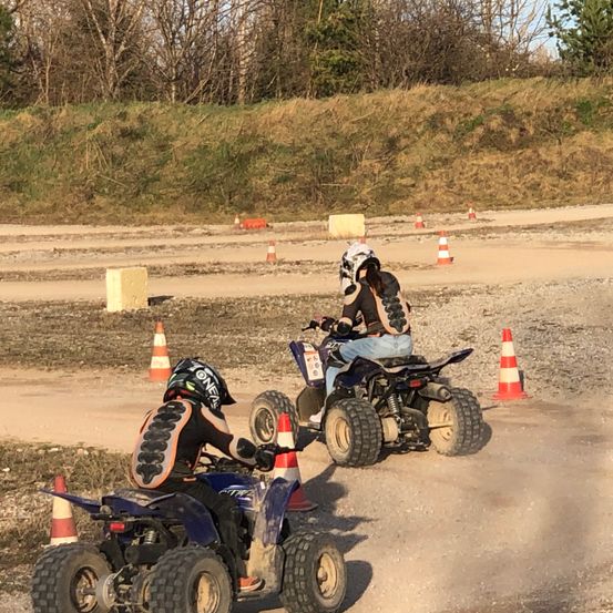 Zwei Personen fahren ATVs auf einer Schotterpiste, umgeben von orangefarbenen Verkehrskegel. Einer trägt einen weißen Helm, der andere einen schwarzen. Die Strecke wird von einem Graswall gesäumt.