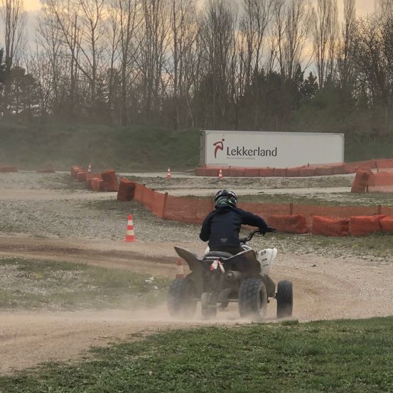 Eine Person fährt ein vierrädriges Fahrzeug auf einer Schotterpiste. Im Hintergrund steht ein Lekkerland-Schild. Das Gebiet ist mit orangefarbenen Barrieren eingezäunt.