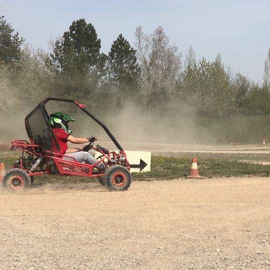 Eine Person mit Helm fährt einen roten Go-Kart auf einer Schotterpiste, mit Bäumen und orangefarbenen Verkehrskegel im Hintergrund.