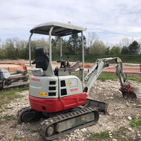 Ein rot-weißer Minibagger steht auf einer felsigen Oberfläche, mit Bäumen und einer Rasenfläche im Hintergrund.