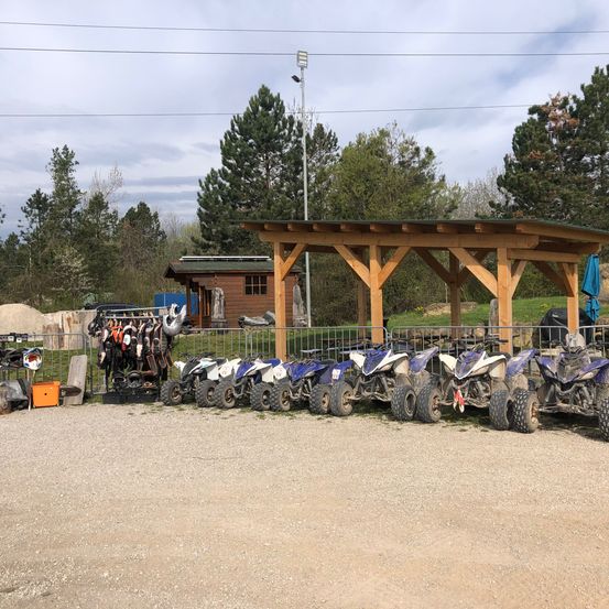 Mehrere ATVs sind unter einem Holzpavillon geparkt. In der Nähe hängen Helme. Der Bereich ist von einem Zaun und Bäumen umgeben. Ein kleines Gebäude ist im Hintergrund zu sehen.