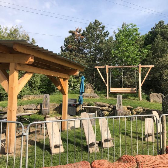Ein hölzerner Pavillon mit Tisch und Grill steht neben einer Schaukel und einem Picknickplatz. Bäume und eine Steinmauer sind im Hintergrund.