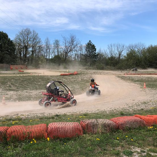 Zwei Personen fahren auf einer Dirt-Strecke mit Go-Karts, mit Bäumen und einem bewölkten Himmel im Hintergrund.