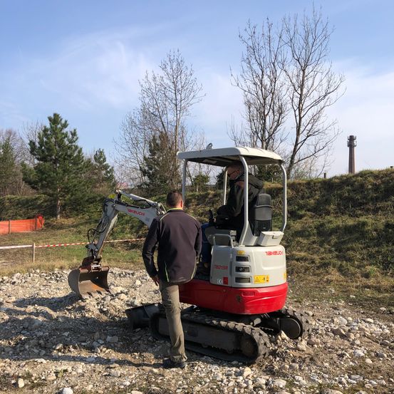 Zwei Männer stehen neben einem roten und weißen Bagger auf einem mit Kies bedeckten Boden, mit Bäumen und einem Turm in der Ferne im Hintergrund.