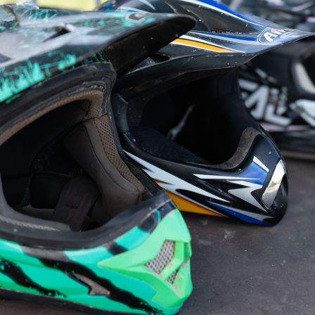 Nahaufnahme von drei Motocross-Helmen. Der vordere ist grün und schwarz mit einem Gittergitter. Dahinter ein weiterer Helm in Blau und Schwarz mit weißen Akzenten. Weiter hinten ein schwarzer Helm mit weißen und gelben Markierungen.