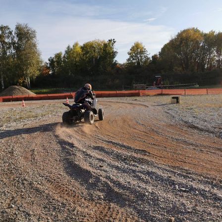 Eine Person fährt einen schwarzen ATV auf einer Erdstrecke, umgeben von Bäumen und orangefarbenen Barrieren. Der Himmel ist klar und blau.