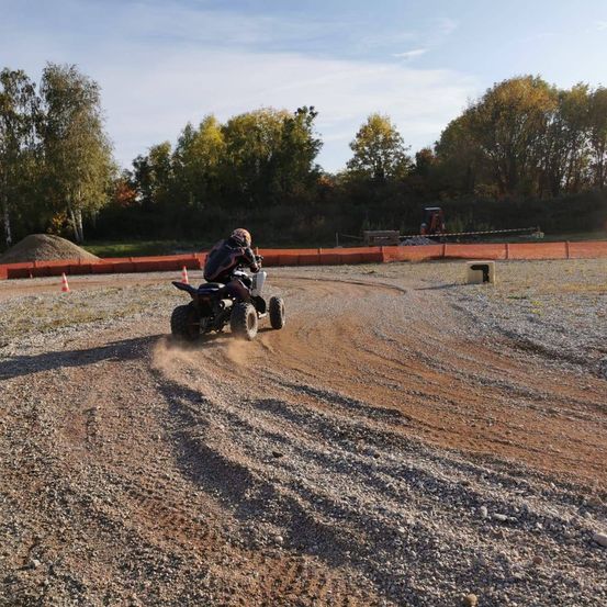 Eine Person fährt einen schwarzen ATV auf einer Erdstrecke, umgeben von Bäumen und orangefarbenen Barrieren. Der Himmel ist klar und blau.