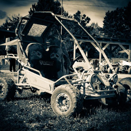 Ein schwarz-weißes Foto zeigt einen Go-Kart auf einem Grasfeld. Er hat ein Frontgitter und ein Verdeck sowie zwei Sitze. Die Umgebung umfasst Bäume und eine Holzkonstruktion.