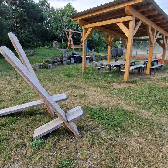 Eine hölzerne Außenstruktur mit einem geneigten Dach steht auf einem Grasfeld. Ein Holzstuhl steht vor dem Boden. Unter der Struktur befinden sich Picknicktische und Bänke.