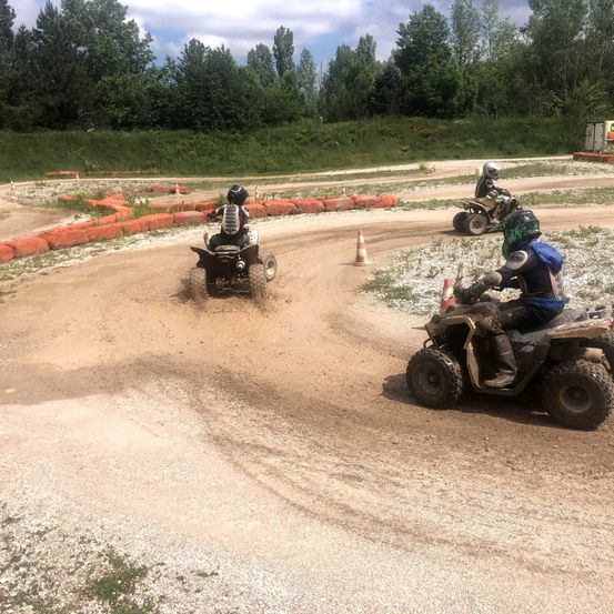 Drei Personen fahren mit ATVs auf einer Erdpiste, eine in Führung, die anderen folgen dicht dahinter. Orangefarbene Barrieren säumen die Strecke, und Bäume und Büsche sind im Hintergrund zu sehen.