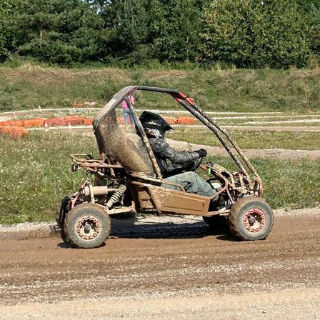 Eine Person fährt einen schmutzigen Go-Kart auf einer unbefestigten Straße. Hinter ihr ist ein Feld mit Gras und Bäumen zu sehen. Eine Person in einem rosa Shirt steht in der Nähe von orangefarbenen Hütchen.