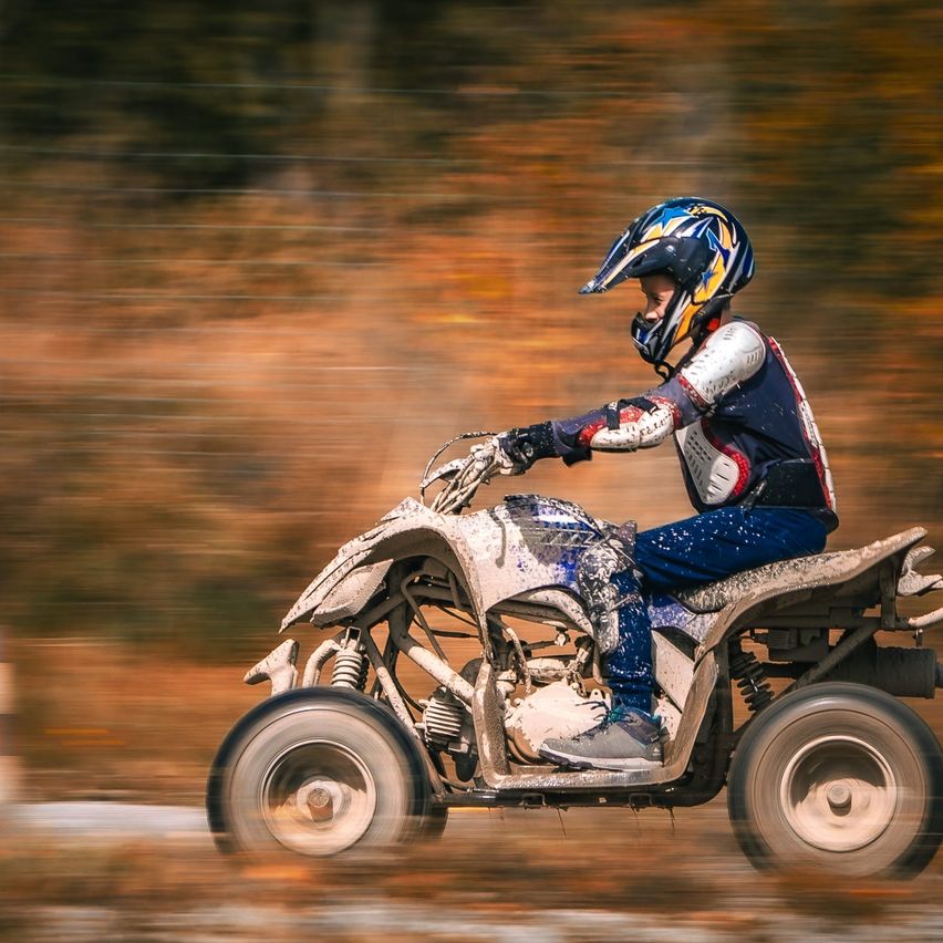 Eine Person mit Helm fährt ein schlammiges Quad auf einem verschwommenen Hintergrund aus Bäumen und Blättern.