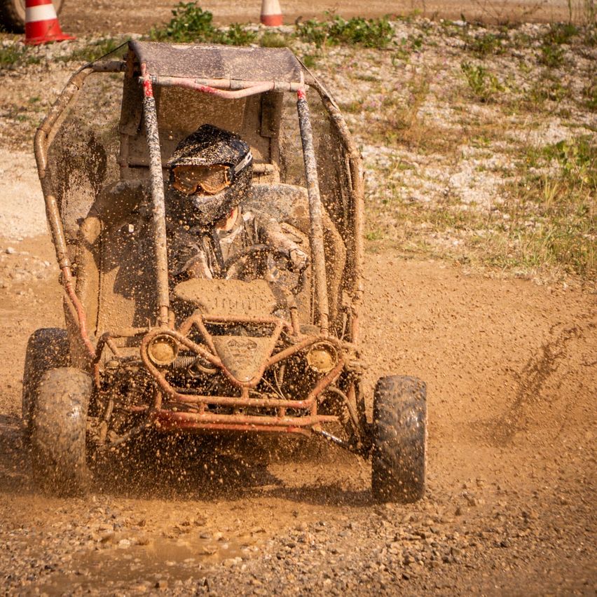 Eine Person fährt einen verschmutzten Go-Kart auf einer Schotterpiste. Der Fahrer trägt einen Helm und eine Schutzbrille. Das Fahrzeug spritzt Schlamm.