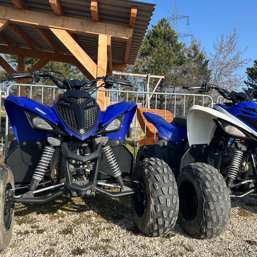 Zwei blaue Yamaha ATVs sind auf Kies geparkt. Sie haben große Reifen und stehen unter einer Holzkonstruktion mit einem Metallzaun. Bäume und ein Strommast sind im Hintergrund.