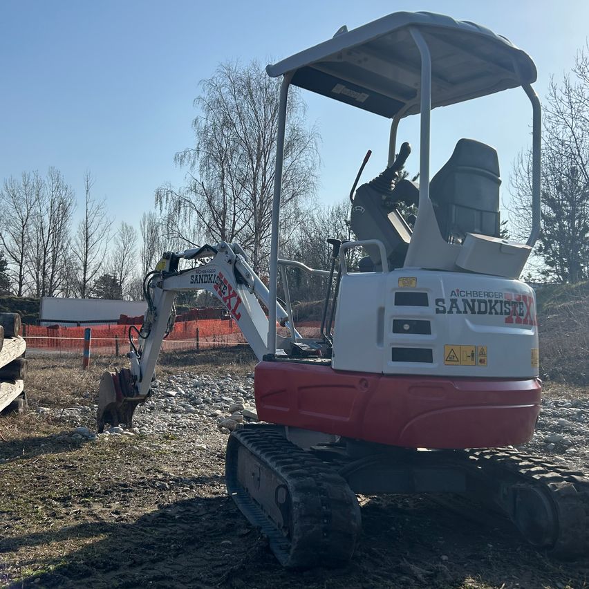 Ein weißer und roter Bagger mit der Aufschrift 'Sandkasten' steht auf einem Erdfeld mit Bäumen und einer Baustelle im Hintergrund.