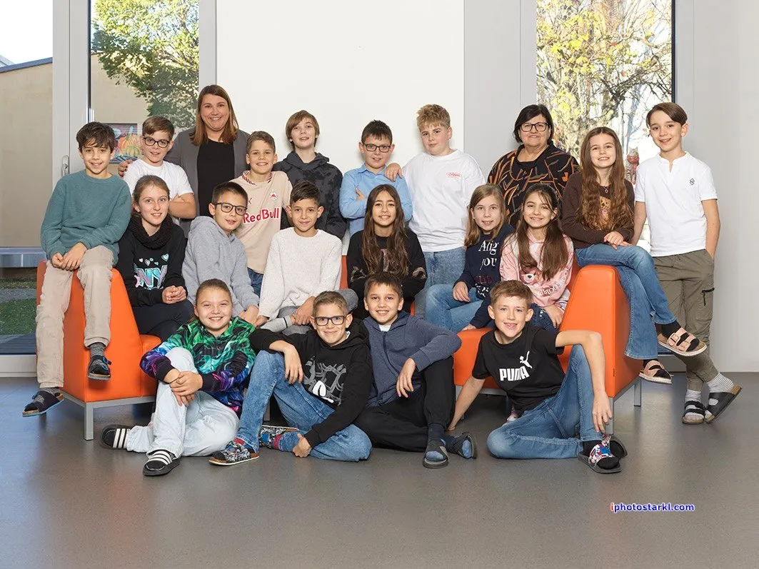 Eine Gruppe von Schülern und zwei Frauen sitzen auf orangefarbenen Sofas im Innenbereich. Die Schüler tragen Freizeitkleidung. Die beiden Frauen tragen Brillen und Jacken.
