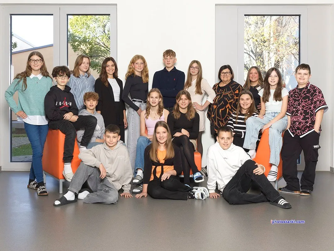 Eine Gruppe von Schülern und Lehrern in einem Raum mit orangefarbenen Stühlen, sitzend und stehend vor einem Fenster.