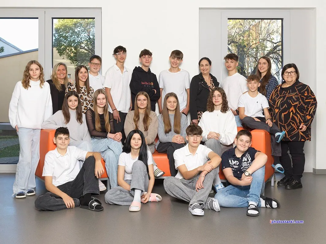Eine Gruppe junger Menschen und Erwachsener posiert für ein Foto. Einige sitzen auf einer orangefarbenen Couch, während andere stehen. Sie befinden sich in einem Raum mit großen Fenstern.