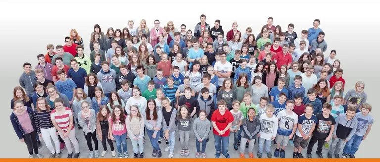 Eine Gruppenaufnahme von Schülern und Lehrern, die auf einem weißen Hintergrund stehen, einige mit Brillen und lässiger Kleidung, lächelnd für die Kamera.