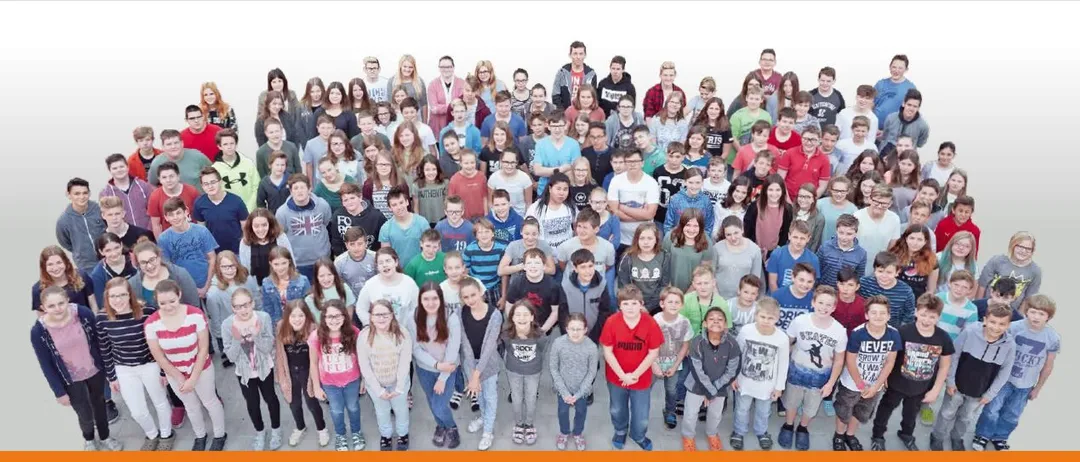 Eine Gruppenaufnahme von Schülern und Lehrern, die auf einem weißen Hintergrund stehen, einige mit Brillen und lässiger Kleidung, lächelnd für die Kamera.