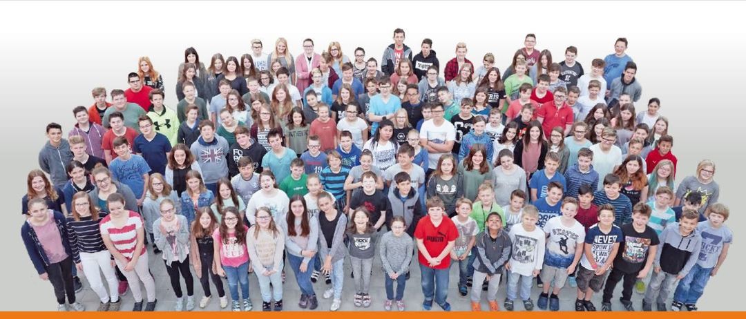 Eine Gruppenaufnahme von Schülern und Lehrern, die auf einem weißen Hintergrund stehen, einige mit Brillen und lässiger Kleidung, lächelnd für die Kamera.