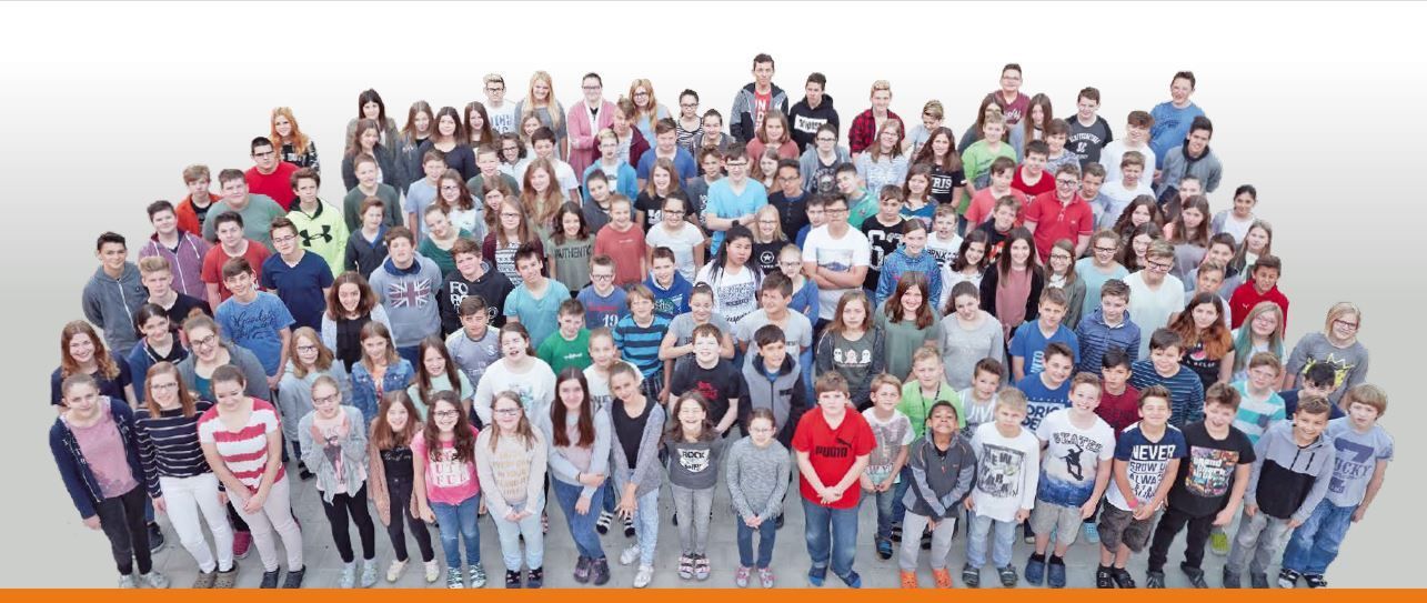 Eine Gruppenaufnahme von Schülern und Lehrern, die auf einem weißen Hintergrund stehen, einige mit Brillen und lässiger Kleidung, lächelnd für die Kamera.