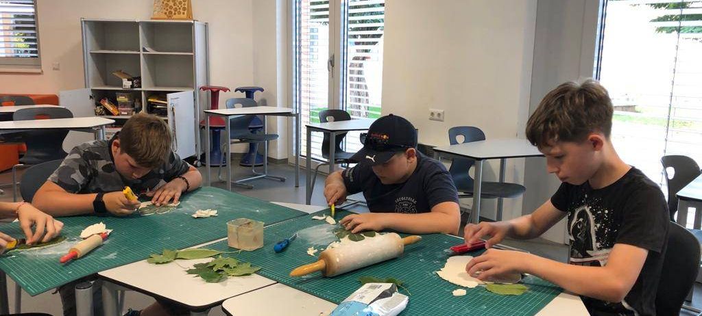 Drei Jungen machen ein Bastelprojekt in einem Klassenzimmer. Sie sitzen an einem Tisch mit einem Nudelholz, Blättern und Werkzeugen. Ein Junge hält ein Messer.