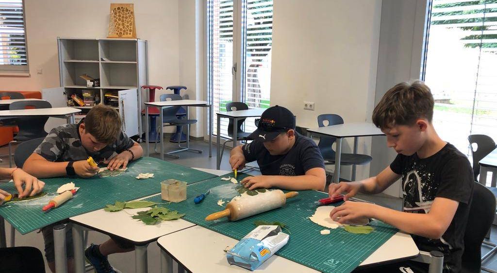 Drei Jungen machen ein Bastelprojekt in einem Klassenzimmer. Sie sitzen an einem Tisch mit einem Nudelholz, Blättern und Werkzeugen. Ein Junge hält ein Messer.