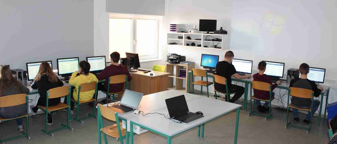 Ein Computerraum mit weißen Wänden, Schüler sitzen an Tischen und nutzen Laptops. Ein Projektor ist an der Decke montiert. Verschiedene elektronische Geräte und ein Drucker sind auf einem Regal.