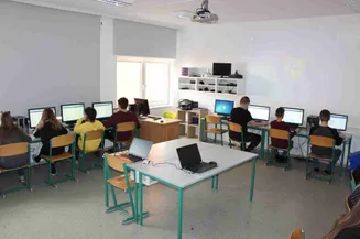 Ein Klassenzimmer mit weißen Wänden, einem Projektor und mehreren Tischen. Schüler sitzen an den Tischen und benutzen Laptops und Monitore. Ein Drucker steht auf dem Tisch.