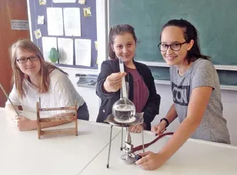 Drei Mädchen in einem Klassenzimmer führen ein wissenschaftliches Experiment durch. Ein Mädchen hält eine Flasche mit Flüssigkeit, ein anderes justiert ein Reagenzglas und das dritte Mädchen beobachtet.