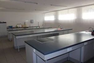 Ein leerer Laborraum mit grauen Arbeitsflächen, weißen Schränken und Metallbeinen. Es gibt Waschbecken, Wasserhähne und Steckdosen. Der Raum hat weiße Wände und geflieste Böden.