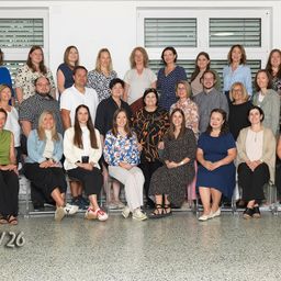 Ein Gruppenfoto von 26 Personen in einem Raum, in Reihen sitzend, einige stehend. Sie lächeln für die Kamera, mit verschiedenen Kleidungsstilen.