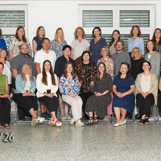 Ein Gruppenfoto von 26 Personen in einem Raum, in Reihen sitzend, einige stehend. Sie lächeln für die Kamera, mit verschiedenen Kleidungsstilen.
